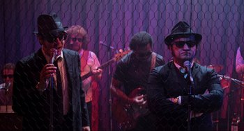 Movie still from “The Blues Brothers” (1980), directed by John Landis – A group of people that are standing in front of a chain link fence; Wide shot, Low angle