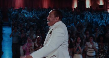 Movie still from “The Blues Brothers” (1980), directed by John Landis – A man in a white suit is singing in front of an audience; Medium shot, Over the shoulder angle