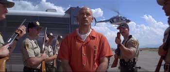 Movie still from “Con Air” (1997), directed by Simon West – A man in an orange prison uniform standing in front of a helicopter; Medium shot, Low angle