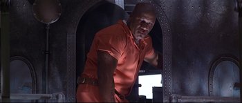 Movie still from “Con Air” (1997), directed by Simon West – A man in an orange prison uniform standing inside a building; Medium shot, Low angle