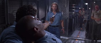 Movie still from “Con Air” (1997), directed by Simon West – A man in blue shirt holding a cell phone in front of another man; Medium shot, Low angle