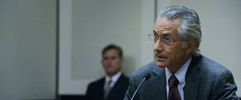 Movie still from “The Bourne Legacy” (2012), directed by Tony Gilroy – A man in a suit and glasses is giving a speech; Close Up shot, Over the shoulder angle