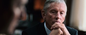 Movie still from “The Bourne Ultimatum” (2007), directed by Paul Greengrass – An older man in a black suit and white shirt is holding his hands under his chin; Close Up shot, Over the shoulder angle