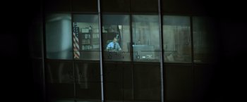 Movie still from “The Bourne Ultimatum” (2007), directed by Paul Greengrass – A man sitting at a desk in front of an american flag reflected in a window; Extreme Wide shot, High angle