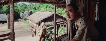 Movie still from “The Bridge on the River Kwai” (1957), directed by David Lean – A man standing in front of a hut on the side of a river; Medium shot, Low angle