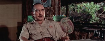 Movie still from “The Bridge on the River Kwai” (1957), directed by David Lean – A man holding a fan while sitting on a chair; Medium shot, Low angle
