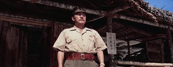 Movie still from “The Bridge on the River Kwai” (1957), directed by David Lean – An older man wearing a hat standing in front of a building; Medium shot, Low angle
