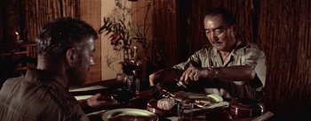Movie still from “The Bridge on the River Kwai” (1957), directed by David Lean – A man sitting at a dinner table with a bottle of wine; Medium shot, Low angle