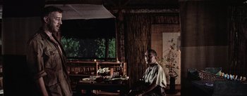 Movie still from “The Bridge on the River Kwai” (1957), directed by David Lean – A man sitting at a table in front of an open window; Wide shot, Low angle
