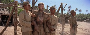 Movie still from “The Bridge on the River Kwai” (1957), directed by David Lean – A group of men standing next to each other on top of a dirt field; Medium shot, Low angle