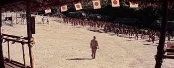 Movie still from “The Bridge on the River Kwai” (1957), directed by David Lean – A man walking down a dirt road in front of a group of soldiers; Extreme Wide shot, High angle