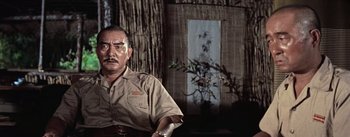 Movie still from “The Bridge on the River Kwai” (1957), directed by David Lean – An older man sitting in front of a tree; Medium shot, Low angle