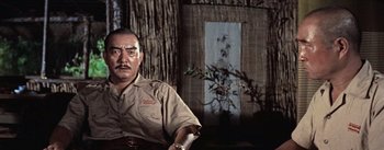 Movie still from “The Bridge on the River Kwai” (1957), directed by David Lean – A man sitting in front of a tree in front of a building; Medium shot, Low angle