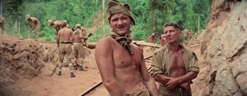 Movie still from “The Bridge on the River Kwai” (1957), directed by David Lean – A man with a bandana on his head is posing for a picture; Medium shot, Low angle
