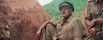 Movie still from “The Bridge on the River Kwai” (1957), directed by David Lean – A man sitting on the side of a hill wearing a hat; Medium shot, Low angle