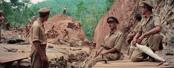 Movie still from “The Bridge on the River Kwai” (1957), directed by David Lean – A man in a uniform sitting on a wooden platform; Wide shot, Low angle