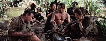 Movie still from “The Bridge on the River Kwai” (1957), directed by David Lean – A group of men and women sitting on the ground in the woods; Medium shot, Over the shoulder angle