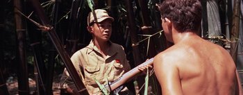 Movie still from “The Bridge on the River Kwai” (1957), directed by David Lean – A man holding a rifle in his hand while standing next to another man; Medium shot, Over the shoulder angle