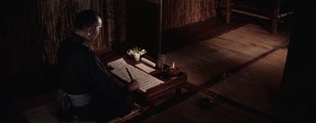 Movie still from “The Bridge on the River Kwai” (1957), directed by David Lean – A man sitting in front of a table with a sheet of music on top of it; Medium shot, High angle