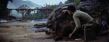 Movie still from “The Bridge on the River Kwai” (1957), directed by David Lean – A man kneeling on the ground next to a large rock; Wide shot, Low angle