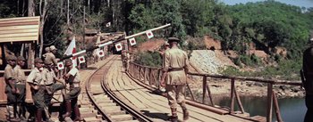 Movie still from “The Bridge on the River Kwai” (1957), directed by David Lean – A man walking across a wooden bridge with trees in the background; Wide shot, High angle
