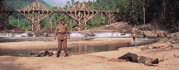 Movie still from “The Bridge on the River Kwai” (1957), directed by David Lean – A man in uniform standing next to a body of water near a bridge; Wide shot, Low angle