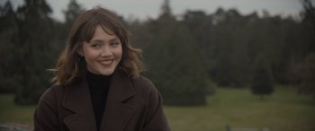 Movie still from “The Bubble” (2022), directed by Judd Apatow – A woman in a brown coat smiling for the camera; Close Up shot, Over the shoulder angle