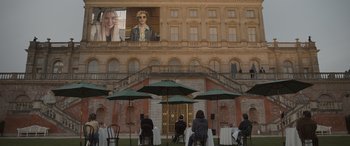 Movie still from “The Bubble” (2022), directed by Judd Apatow – People sitting at tables with umbrellas in front of a building; Extreme Wide shot, Low angle