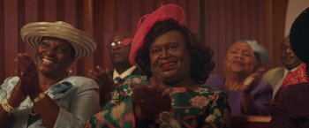 Movie still from “The Burial” (2023), directed by Maggie Betts – An older woman wearing a pink hat clapping her hands; Close Up shot, Over the shoulder angle