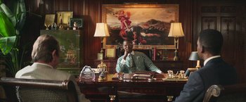 Movie still from “The Burial” (2023), directed by Maggie Betts – A man sitting at a desk in front of a painting on the wall; Medium shot, Over the shoulder angle