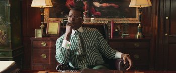 Movie still from “The Burial” (2023), directed by Maggie Betts – A man sitting in a chair in front of a painting; Medium shot, Low angle