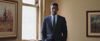 Movie still from “The Burial” (2023), directed by Maggie Betts – A man in a suit and tie standing in front of a window; Medium shot, Low angle