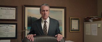 Movie still from “The Burial” (2023), directed by Maggie Betts – A man in a suit and tie sitting in a chair in front of a painting; Medium shot, Low angle