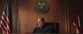 Movie still from “The Burial” (2023), directed by Maggie Betts – A judge sitting in front of a judge's chair in front of a seal of the u; Medium shot, Low angle