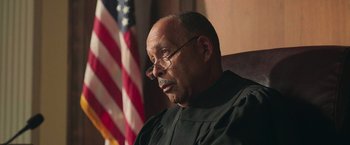 Movie still from “The Burial” (2023), directed by Maggie Betts – A man wearing a black robe and glasses in front of an american flag; Close Up shot, Low angle