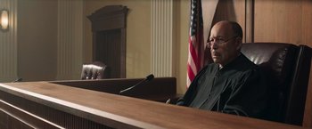 Movie still from “The Burial” (2023), directed by Maggie Betts – A man sitting in front of a microphone in a courtroom; Medium shot, Low angle