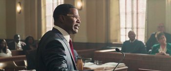 Movie still from “The Burial” (2023), directed by Maggie Betts – A man in a business suit is speaking at a podium; Medium shot, Over the shoulder angle