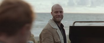 Movie still from “The Burning Sea” (2021), directed by John Andreas Andersen – A man with a shaved head smiling for the camera; Close Up shot, Over the shoulder angle