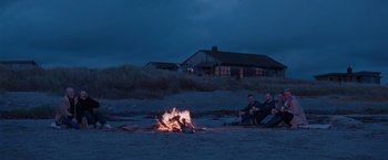 Movie still from “The Burning Sea” (2021), directed by John Andreas Andersen – A couple of people sitting around a fire on the beach; Wide shot, High angle