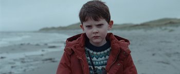 Movie still from “The Burning Sea” (2021), directed by John Andreas Andersen – A young boy in a red jacket standing on the beach; Close Up shot, Over the shoulder angle