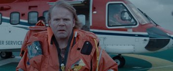 Movie still from “The Burning Sea” (2021), directed by John Andreas Andersen – A man in an orange jacket standing next to a helicopter; Medium shot, Over the shoulder angle