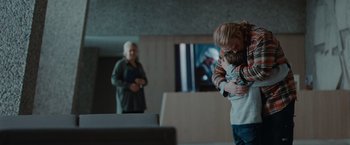 Movie still from “The Burning Sea” (2021), directed by John Andreas Andersen – Two people hugging in a room with a television in the background; Medium shot, Over the shoulder angle