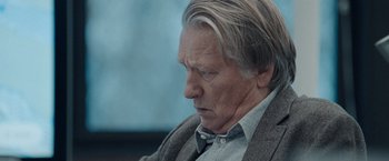 Movie still from “The Burning Sea” (2021), directed by John Andreas Andersen – An older man with a gray shirt and a gray jacket; Close Up shot, Over the shoulder angle