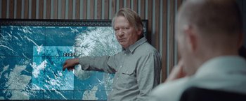 Movie still from “The Burning Sea” (2021), directed by John Andreas Andersen – A man standing in front of a wall with a map on it; Medium shot, Over the shoulder angle