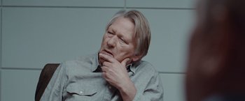Movie still from “The Burning Sea” (2021), directed by John Andreas Andersen – An older man with long blonde hair is holding his chin in his hand; Close Up shot, Low angle