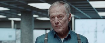 Movie still from “The Burning Sea” (2021), directed by John Andreas Andersen – An older man wearing a blue shirt and a black bag; Close Up shot, Low angle