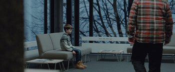 Movie still from “The Burning Sea” (2021), directed by John Andreas Andersen – A young boy sitting on a couch in a room; Wide shot, Over the shoulder angle