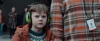 Movie still from “The Burning Sea” (2021), directed by John Andreas Andersen – A young boy wearing ear muffs and a plaid jacket; Close Up shot, Over the shoulder angle