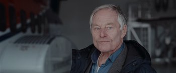 Movie still from “The Burning Sea” (2021), directed by John Andreas Andersen – An older man wearing a blue shirt and a jacket; Close Up shot, Low angle