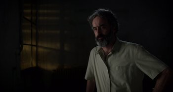 Movie still from “Crimes of the Future” (2022), directed by David Cronenberg – A man with a gray beard is standing in a dark room; Medium shot, Low angle